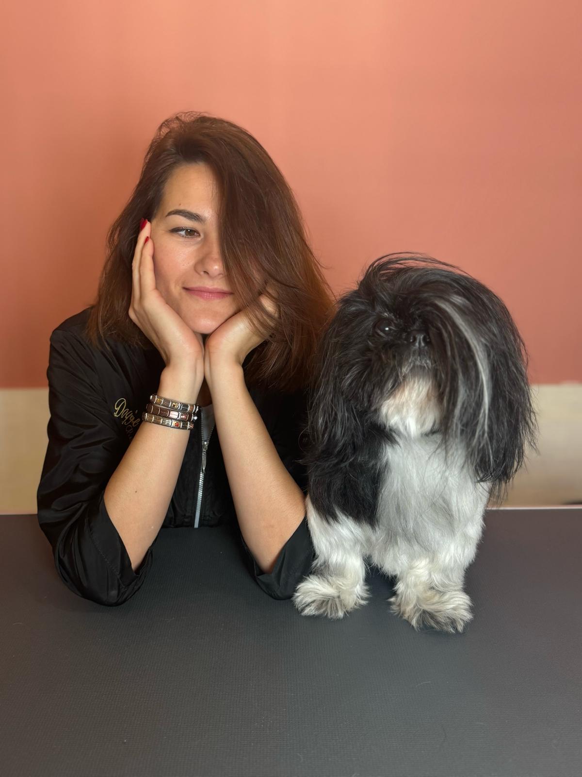 Kobieta z długimi brązowymi włosami opiera głowę na dłoniach i siedzi obok czarno-białego psa rasy Shih Tzu na tle brzoskwiniowej ściany.